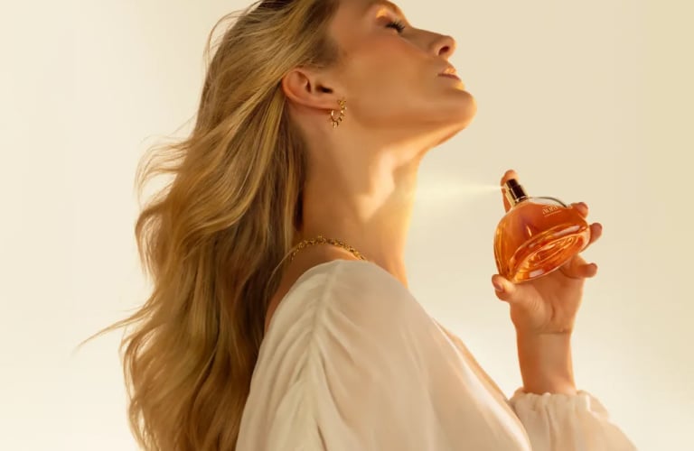 Foto com fundo claro de uma mulher loira vestindo blusa branca, de perfil e com a cabeça inclinada para cima, borrifando um perfume arredondado no pescoço.